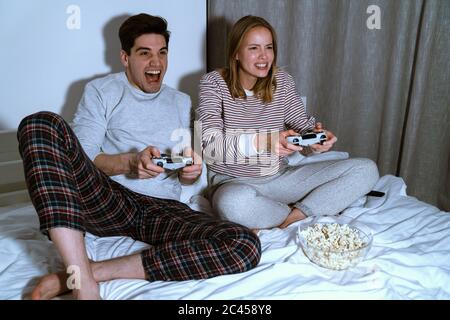 Portrait von aufgeregt niedlichen Paar hält Joysticks während der Wiedergabe von Videospielen im Bett zu Hause Stockfoto