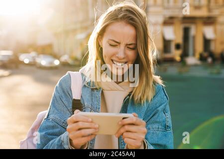 Schöne verwirrt junge blonde Frau trägt Denim Jacke zu Fuß in der Stadt, mit Handy Stockfoto