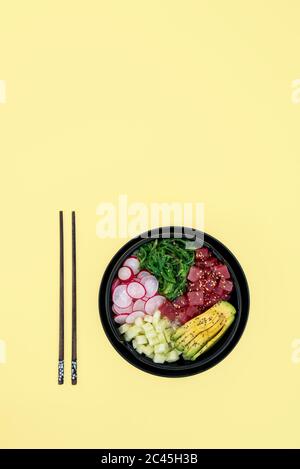 Draufsicht auf Thunfisch-Poke-Schüssel mit Reis, Avocado, Wakame, Rettich und Gurke auf gelbem Hintergrund Stockfoto
