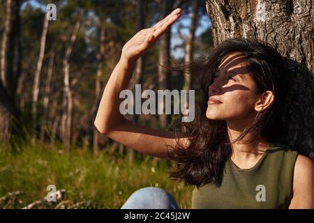 Porträt einer jungen Frau, die unter einem Baum in einem Wald sitzt und ihre Augen vor der Sonne beschattete Stockfoto