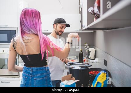 Junge Frau mit langen rosa Haaren und bärtigen Mann mit Baseball-Mütze in einer Küche stehend, Kochen. Stockfoto