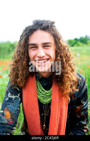 Porträt eines jungen Mannes mit langen braunen lockigen Haaren auf einer Mohn-Wiese, lächelnd an der Kamera. Stockfoto