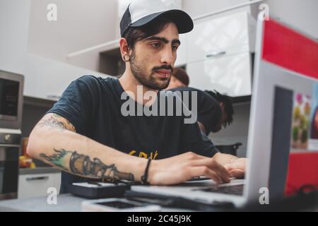 Junger bärtiger Mann mit Piercings, der an einem Tisch sitzt, Baseballmütze trägt und auf dem Laptop tippt. Stockfoto