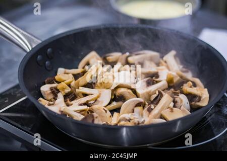 Prozess der Zubereitung von Champignon-Champignons in Scheiben geschnitten in schwarzer Pfanne mit Öl auf Elektroherd. Professionelle Küche, Catering, Kochen, Gastronomie und Stockfoto