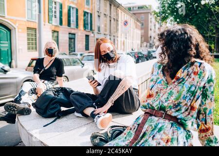 Drei junge Frauen tragen Gesichtsmasken während des Corona-Virus, sitzen an einem Flussufer, plaudern. Stockfoto