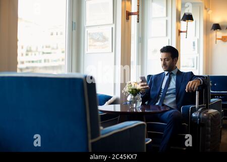 Geschäftsmann, der mit Koffer in der Hotellobby sitzt und das Handy benutzt. Stockfoto