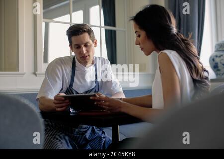 Chefkoch mit blauer Schürze und Frau, die an einem Tisch sitzt und auf ein digitales Tablet schaut. Stockfoto