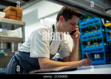 Koch trägt blaue Schürze in der Küche stehend, mit Handy. Stockfoto