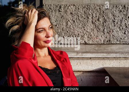 Porträt einer sitzenden Frau mit langen Brünetten Haaren, trägt rote Jacke, Hand in Haar. Stockfoto