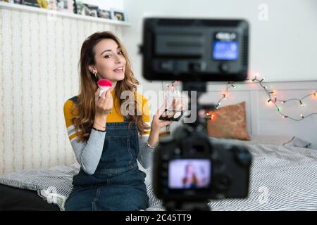 Junge Frau mit langen braunen Haaren auf dem Bett sitzend, hält Make-up und Pinsel, Aufnahme Blog. Stockfoto