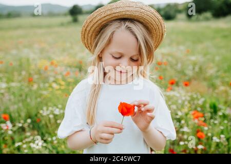 Portrait eines jungen Mädchens, das einen Mohn hält, auf einer Wiese voller wilder Blumen stehend. Stockfoto