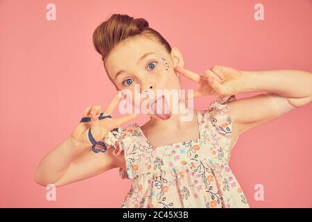 Portrait von Brünette Mädchen tragen Rüschen Kleid mit Blumenmuster auf rosa Hintergrund, herausragend Zunge bei der Kamera. Stockfoto