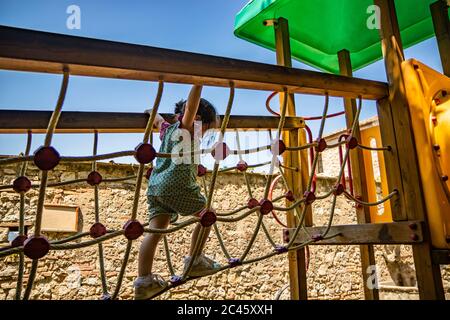 Ein kleines Mädchen spielt gerne auf einer tibetischen Brücke, aus Seil, auf einem Spielplatz mit Rutsche und Rohr. Ein Spiel eines öffentlichen Parks in Narni, Umbrien, Ita Stockfoto