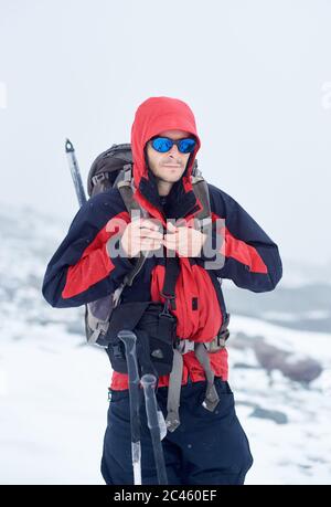 Männliche Tourist in Sonnenbrille Befestigung Reise Rucksack und Blick auf Kamera. Wanderer mit Umhängetasche trägt Sport-Winterjacke mit Kapuze und modischen Farbtönen. Konzept von Reisen, Wandern und Alpinismus Stockfoto