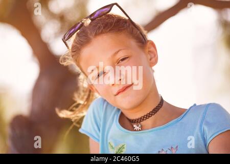 Junges Mädchen, zwischen Alter, auf einem Baum sitzend, lächelnd an Kamera mit Sonnenbrille über Kopf Stockfoto