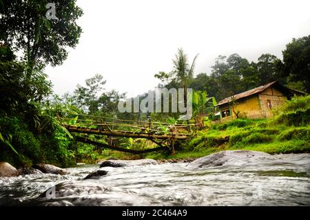 Tropische Landschaft mit felsigen Fluss unter der Holzbrücke und typisch ländliches indonesisches Haus im Dschungel von West-Java Stockfoto