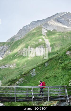 Seitenansicht der Dame Reisende mit Rucksack zu Fuß auf der Brücke mit grasbewachsenen Hügeln im Hintergrund. Junge Frau mit Wanderstöcken wandern allein in den Bergen. Konzept von Reisen, Wandern und Tourismus. Stockfoto