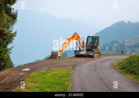 Ein orangefarbener Bagger baut eine alpine Straße in großer Höhe. Die landschaftlich reizvolle Bergstraße in den italienischen Alpen wird im Sommer, Lombardei, Italien, saniert. Stockfoto