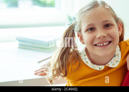 Portrait of adorable teen Mädchen mit blonden Zöpfen und niedlichen Lächeln auf dem Schulschreibtisch mit Büchern im Hintergrund Stockfoto