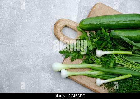 Holzschneidebrett mit frischen grünen Zwiebeln, Gurken, Petersilie, Dill auf Steingrund. Draufsicht. Stockfoto