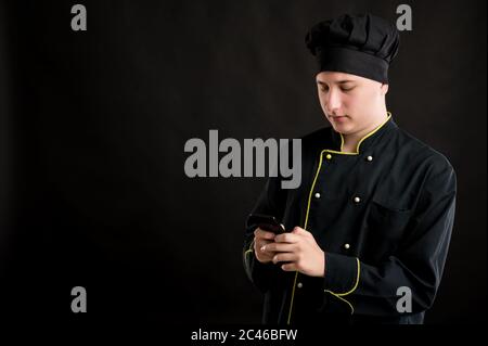 Portrait von jungen Mann in einem schwarzen Koch Anzug mit blonden Haaren gekleidet, Lesen und Senden von SMS auf Handy posiert auf einem schwarzen isolierten Backgr Stockfoto