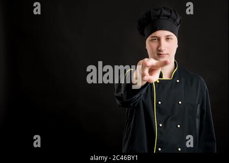 Portrait von jungen Mann in einem schwarzen Koch Anzug gekleidet tun Ich beobachte Sie Geste posiert auf einem schwarzen isolierten Hintergrund mit Kopie Raum Werbung Stockfoto