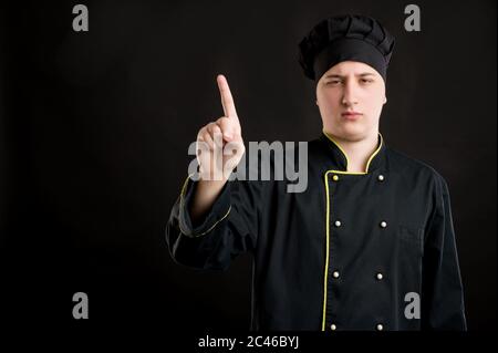 Portrait von jungen Mann in einem schwarzen Koch Anzug gekleidet zeigt Nein mit ihrem Finger posiert auf einem schwarzen isolierten Hintergrund mit Kopie Raum Werbefläche Stockfoto