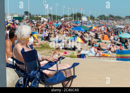 Littlehampton, Sussex, Großbritannien 24. Juni 2020. UK Weather - eine ältere Frau mit Gesichtsmaske hält ihre Distanz und sitzt weg von den Massen von Menschen, die am Strand sind und das sonnige Wetter genießen. Quelle: Scott Ramsey/Alamy Live News. Stockfoto