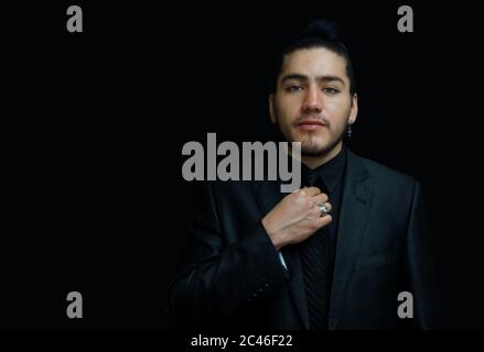 Porträt eines jungen lateiner mit Ohrring mit schwarzem Anzug, schwarzem Hemd und schwarzer Krawatte mit der Hand auf der Krawatte von vorne auf schwarzem Hintergrund gesehen Stockfoto