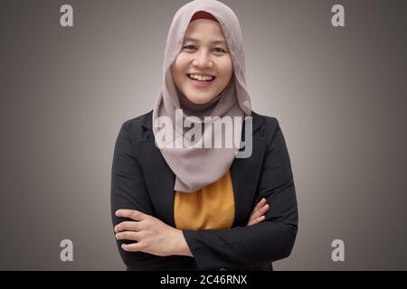 Asiatische muslimische Geschäftsfrau trägt Hijab freundlich lächelnd mit gekreuzten Armen; erfolgreiche selbstbewusste junge Frau; vor grauem Hintergrund Stockfoto