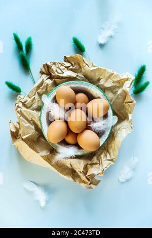 Ein flaches Foto des Osterstilllebens von einer Schale aus braunen Eiern auf zerknittertem Bastelpapier, weißen Federn und trockenen grünen Pflanzen als Dekor. Hellblaue Rückseite Stockfoto