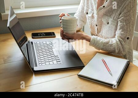 Die Hände der Frau halten eine Tasse, mit Laptop auf dem Schreibtisch in der Wohnung. Remote-Arbeit. Freiberuflich. Ein Smartphone, ein Notebook Stockfoto