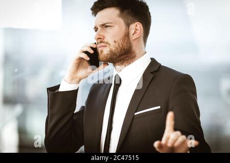 Geschäftskonzept. Junger Geschäftsmann im Büro stehend und beschäftigt am Telefon sprechen zeigt mit der Hand, er löst ein sehr ernstes Arbeitsproblem Stockfoto
