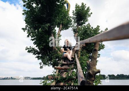 Eine junge Frau mit einem drei Monate alten schottischen Straight Kitten sitzt im Sommer auf einem schönen grünen Baum über dem See. Gehen, ruhen Sie mit einem Haustier. Die Blondine trägt kurze Denim-Shorts, ein schwarzes Baumwoll-T-Shirt und eine Brille. Stockfoto