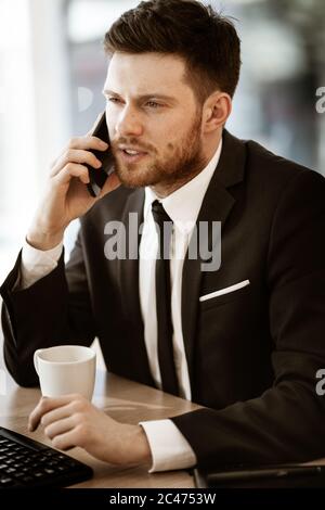 Krisenkonzept. Junger Geschäftsmann am Bürotisch sitzen beschäftigt Gespräch auf einem Handy Lösung eines sehr ernsten Arbeitsproblem. Mann in Anzug Stockfoto
