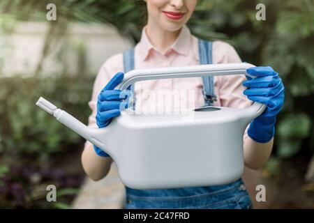 Zugeschnittenes Bild von hübschen weiblichen Floristen in Jeans Overalls und blauen Gummihandschuhe, hält graue Wasserkanne für die Bewässerung von Pflanzen in ihrem Gewächshaus. Stockfoto