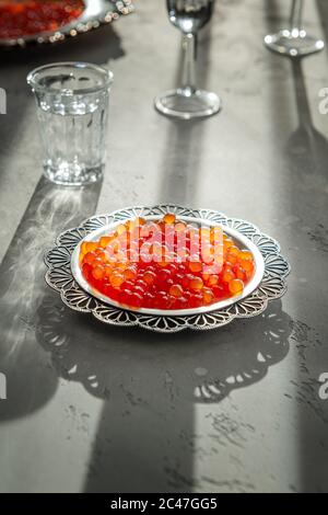 Natürlicher roter Kaviar auf silbernem Teller auf grauem Hintergrund. Wodka Schüsse und Teller mit Kaviar auf unfokussierten Hintergrund. Selektiver Fokus. Gesunde Ernährung concep Stockfoto