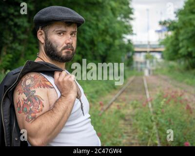 Junger, hübscher, bärtiger Mann mit Tattoos und Kohlestatzen auf der Haut, der auf einer stillstehenden Eisenbahnstrecke mit Blumen mit natürlichen Motiven draußen aufgenommen wurde Stockfoto