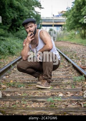 Junger französischer bärtiger Mann, der in Paris, Frankreich, draußen auf einer stillstehenden Eisenbahnstrecke mit natürlichem Licht raucht, mit Tattoos Stockfoto