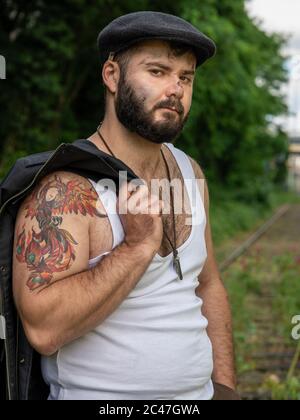 Junger, hübscher, bärtiger Mann mit Tattoos und Kohlestatzen auf der Haut, der auf einer stillstehenden Eisenbahnstrecke mit Blumen mit natürlichen Motiven draußen aufgenommen wurde Stockfoto