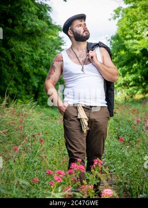 Junger, hübscher, bärtiger Mann mit Tattoos und Kohlestatzen auf der Haut, der auf einer stillstehenden Eisenbahnstrecke mit Blumen mit natürlichen Motiven draußen aufgenommen wurde Stockfoto