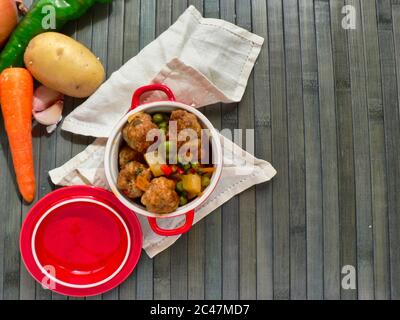 Draufsicht auf einen Topf Frikadellen mit Kartoffeln und Erbsen und rohes Gemüse auf einem Holztisch Stockfoto