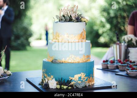 Wunderschöne dreistufige Hochzeitstorte. Hellblauer Kuchen mit goldenem Teller und verschiedenen Blumen verziert. Außenhintergrund. Hochzeitskonzept. Stockfoto