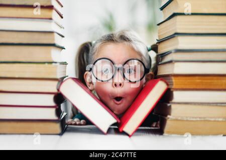 Mädchen im Vorschulalter mit wunderndem Gesicht in großen lustigen Gläsern unter einem Stapel von Büchern Stockfoto