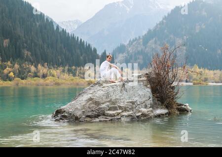 Schönes männliches Modell in weißer Kleidung, das auf einem Felsen sitzt, ein Hintergrund von See und Bergen. Stockfoto
