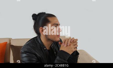 Junger hispanic Mann mit gerafften Haaren gemacht Bogen trägt schwarzes T-Shirt und schwarze Lederjacke sitzen auf einem Sofa, der nach rechts mit Händen zusammen Stockfoto