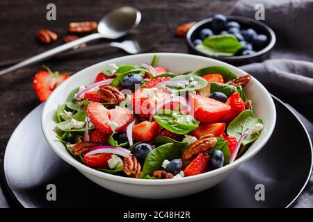 Nahaufnahme von Sommersalat mit Erdbeeren, Heidelbeeren, Spinat, Pekannüssen und zerbröckeltem Feta-Käse in einer weißen Schüssel auf einem dunklen Holztisch, Landschaft Stockfoto