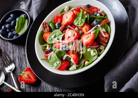 Sommersalat aus Erdbeeren, Blaubeeren, Spinat, Pekannüssen und zerbröckeltem Feta-Käse in einer weißen Schüssel auf einem dunklen Holztisch mit grauem Tuch, Horizont Stockfoto