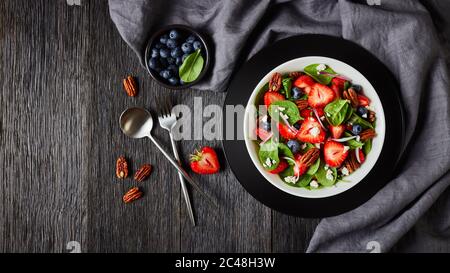 Sommersalat aus Erdbeeren, Blaubeeren, Spinat, Pekannüssen und zerbröckeltem Feta-Käse in einer weißen Schüssel auf einem dunklen Holztisch mit grauem Tuch, Horizont Stockfoto