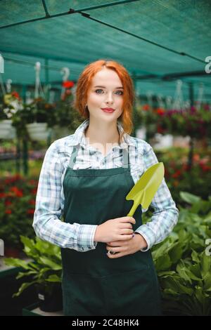 Hübsche Floristin im Schürze stehend mit kleiner Gartenschaufel in der Hand. Junge Rotschopf Dame suchen in der Kamera Stockfoto